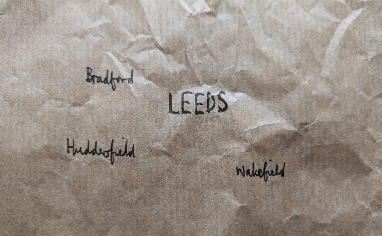 A hand-drawn map of Leeds and towns in vicinity (Bradford, Huddersfield and Wakefield) in black-ink on a brown paper bag showing the West Yorkshire metro public transport network. The text is scaled to the approximate population of the towns in the region.